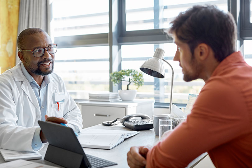 A Black male doctor wearing glasses sitting at a desk across from a young adult male patient discussing the role of testosterone in men’s health