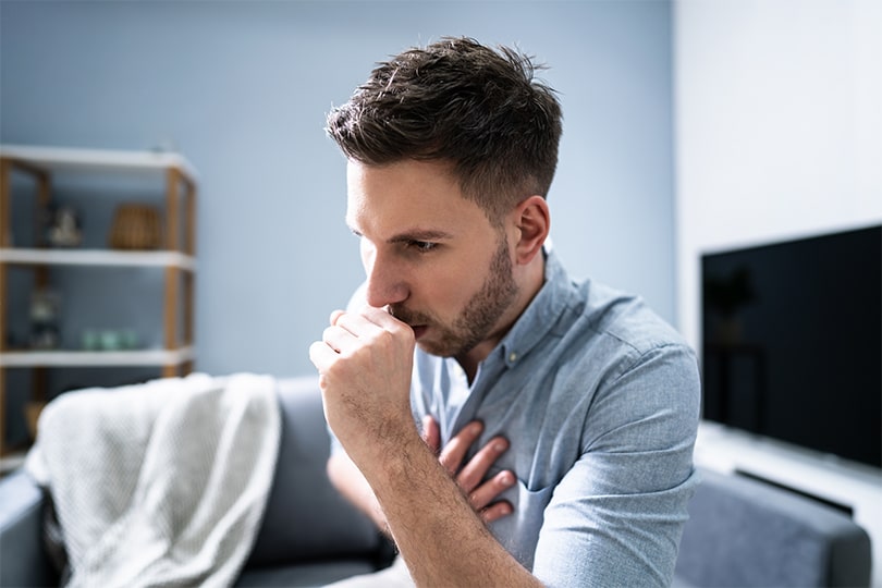 A young adult man coughing and holding his chest, wondering if his symptoms may be a sign of tuberculosis infection