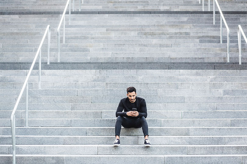 A young adult man sitting outside on a set of steps looking at his phone, reading about the difference between total testosterone and free testosterone.