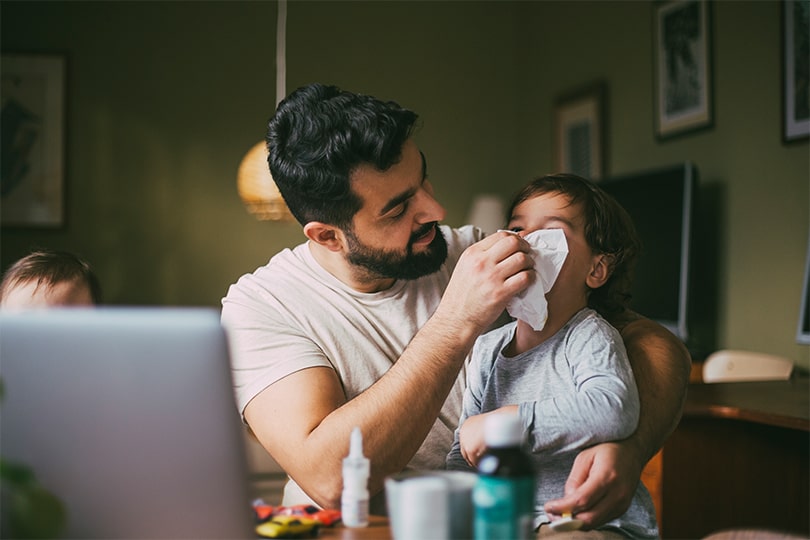 A father sitting with his sick toddler son on his lap, wiping his nose with a tissue and wondering if zinc can help strengthen his immune system and protect him from catching his son’s cold. 