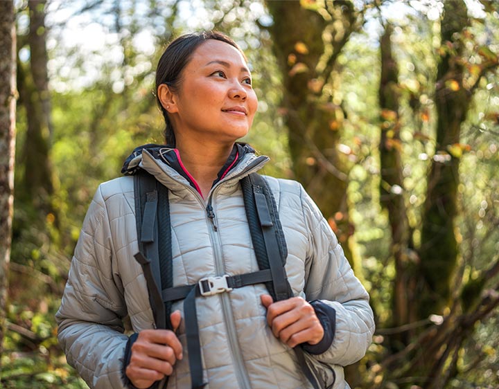 A woman wearing a jacket and carrying a backpack stands amidst a serene forest setting.