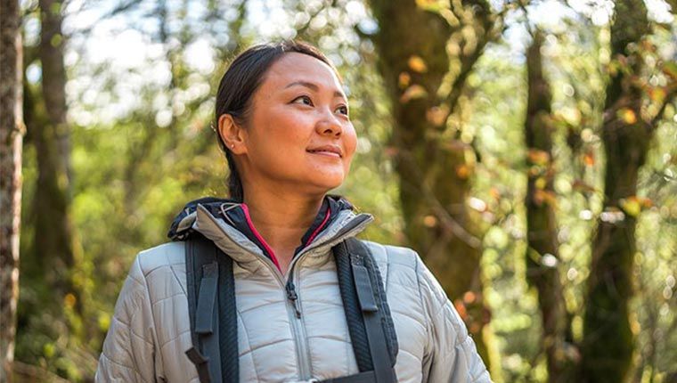 A woman wearing a jacket and carrying a backpack stands amidst a serene forest setting.